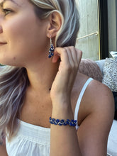 Load image into Gallery viewer, A woman wearing a matching jewelry set featuring a silver-toned bracelet and dangling earrings with dark blue stone-like beads, shown in a close-up lifestyle shot.