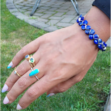 Load image into Gallery viewer, A close-up of a person's hand wearing a blue beaded bracelet and several rings with colorful stones, photographed outdoors on grass and a paved surface.
